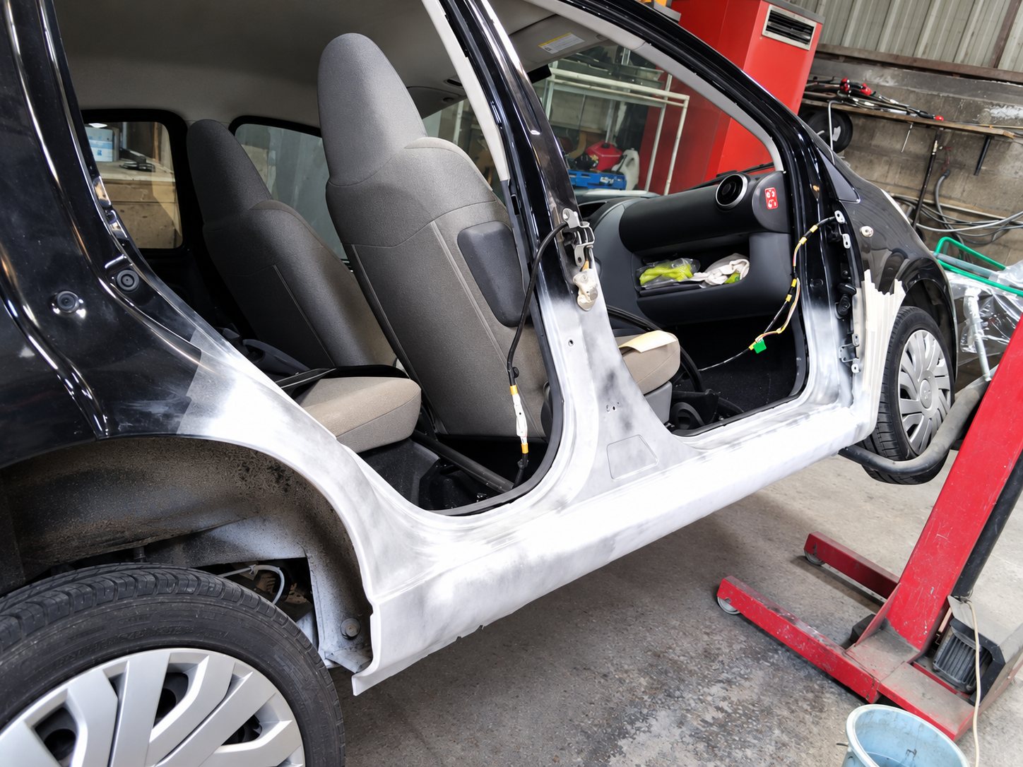 Voiture noire sans portières dont le bas de caisse est poncé et enduit d’apprêt blanc, dans un atelier de carrosserie.