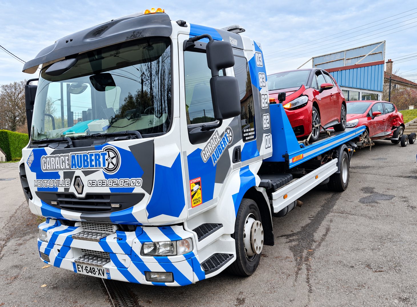 Camion de dépannage blanc et bleu aux couleurs du Garage Aubert, avec le numéro de téléphone sur la cabine, transportant deux voitures rouges accidentées sur son plateau et sa remorque.