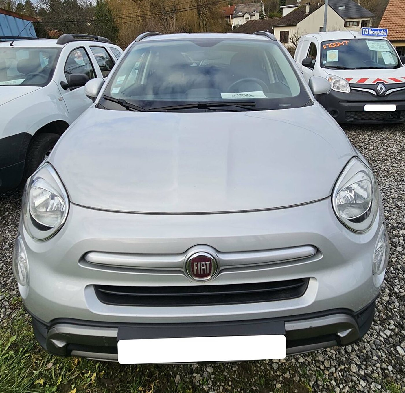 Une Fiat 500X grise vue de face, garée sur le gravier devant le garage.