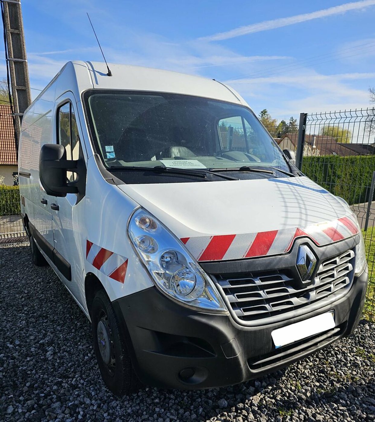 Un fourgon utilitaire Renault blanc orné de bandes de signalisation rouges et blanches, garé sur du gravier devant une clôture et une haie.
