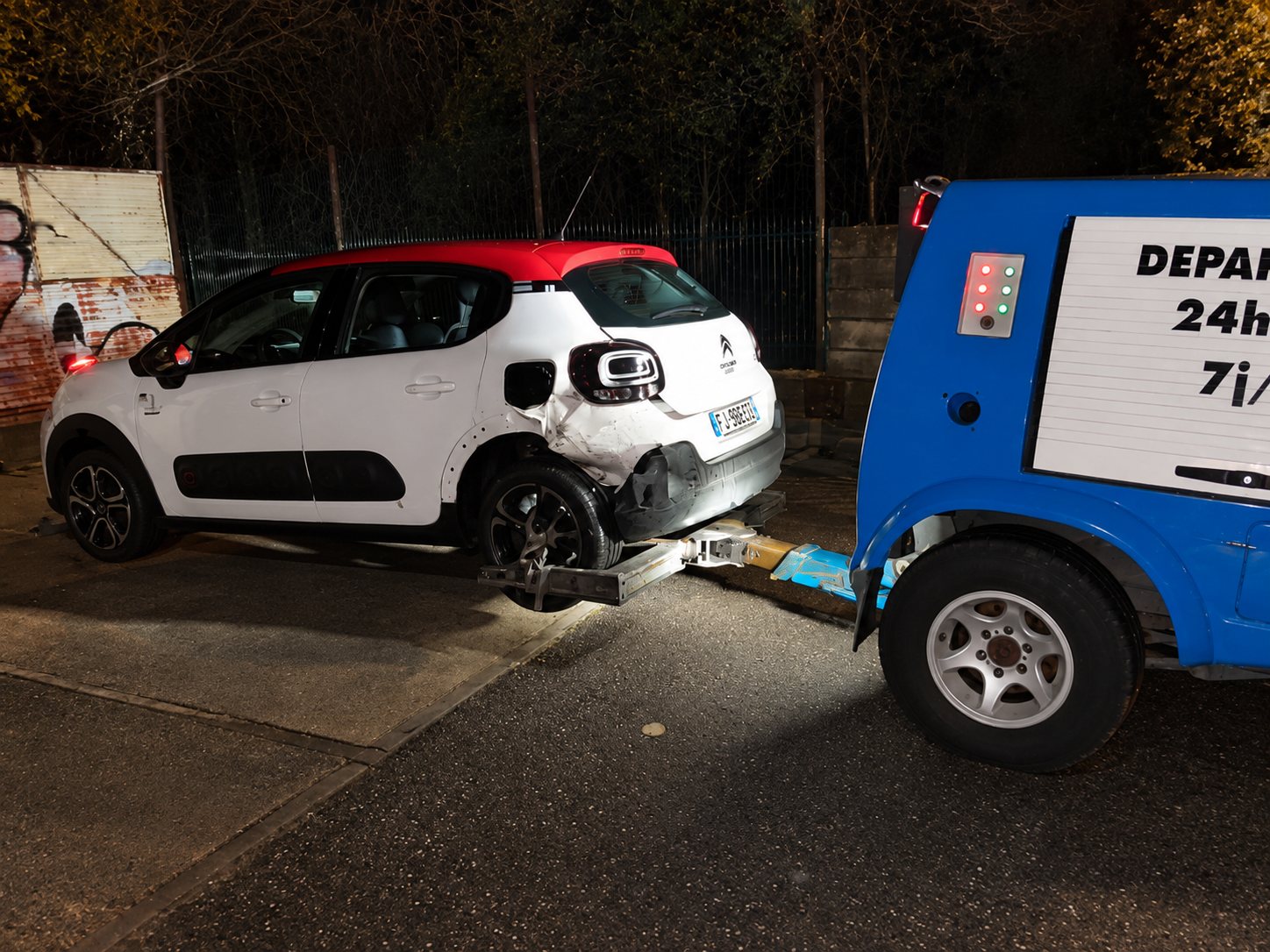 Une Citroën blanche et rouge dont l’arrière est endommagé est remorquée de nuit par une dépanneuse bleue.