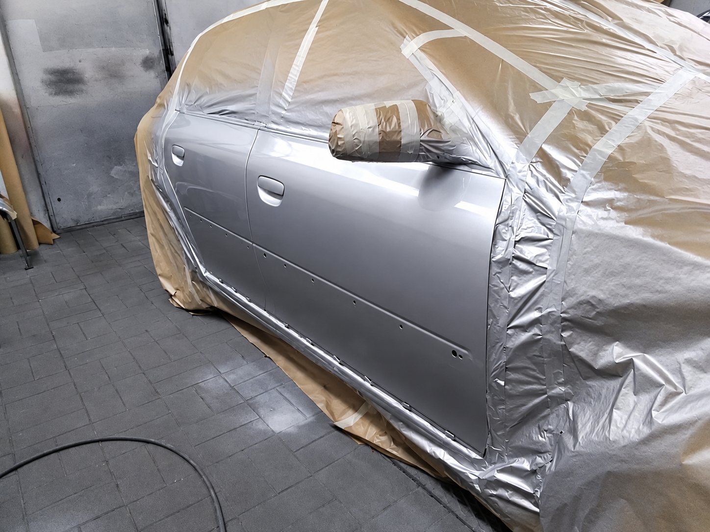 Une voiture argentée dont la portière est fraîchement peinte, le reste de la carrosserie étant recouvert de film plastique et de ruban de masquage dans un atelier.
