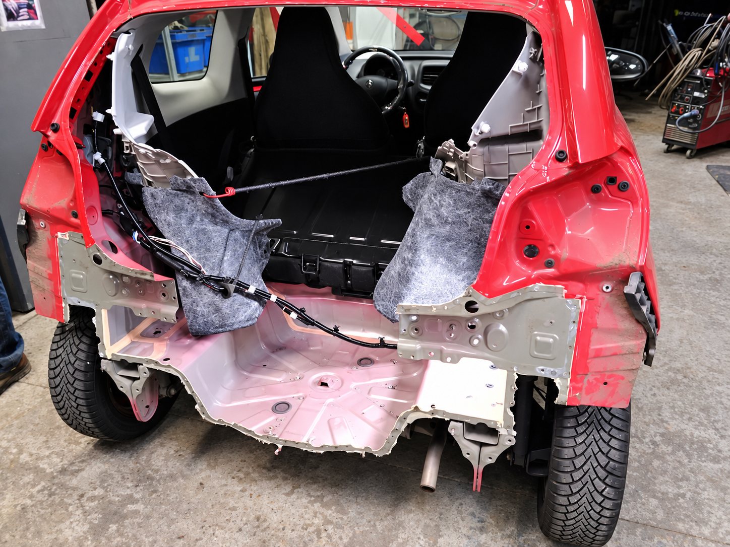 Une petite voiture rouge dans un atelier, dont l’arrière a été découpé et dont la tôle et l’habitacle sont à nu pendant une réparation de carrosserie.